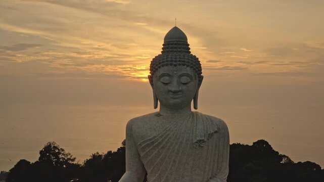 Big Buddha statue sitting peacefully, facing the ocean and lush green hills of Phuket Island in Thailand during tranquil and colorful golden hour sunset. Tourist attraction. Aerial view drone panorama