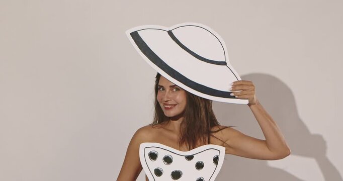 Smiling woman poses with a handmade paper sun hat and matching hand colored top against a neutral studio wall. Concept diy fashion, craft branding and creative editorial content.