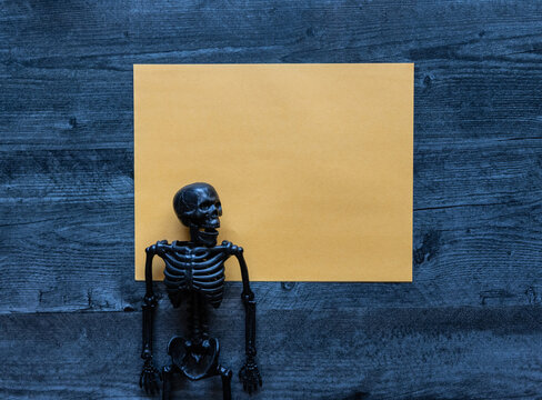 Halloween concept photo of yellow blank paper with a black plastic skeleton flat lay background.