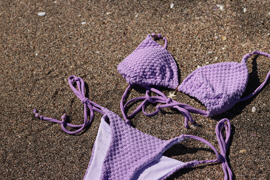 Lilac bikini swimsuit lies on a sandy beach, close-up. Aesthetics of summer vacation. Stylish lilac swimsuit lies on warm sand. Concept of vacation, relaxation by the sea and beach fashion
