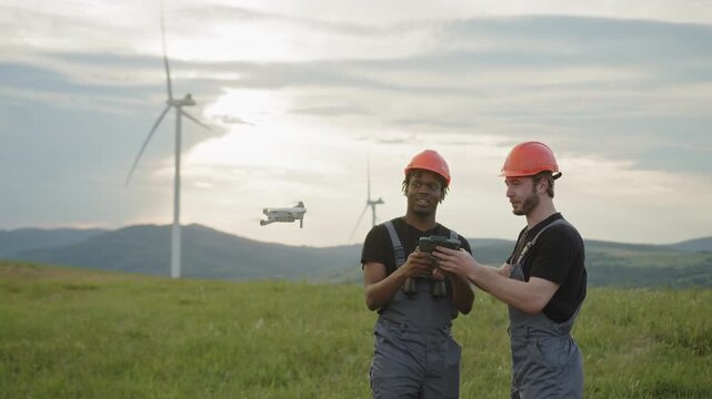 Two engineers in hard hats and overalls stand in a grassy field with wind turbines in the background, one holding binoculars and the other a drone controller