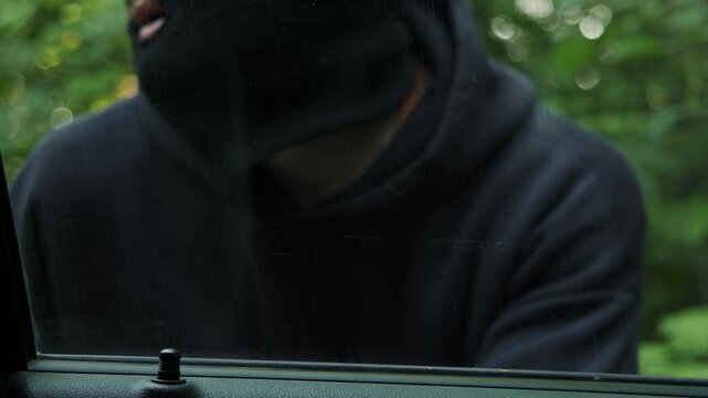 Car thief in balaclava peers through side window while watching road around him. Intruder forces door open and takes control of vehicle to drive away.
