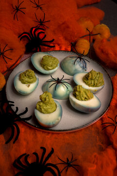 Deviled eggs decorated with green guacamole filling and black spiders on a plate surrounded by orange cobwebs and decorative pumpkins