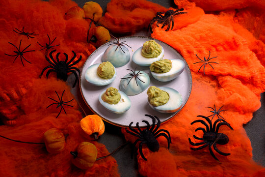 Deviled eggs decorated with green guacamole filling and black spiders on a plate surrounded by orange cobwebs and decorative pumpkins
