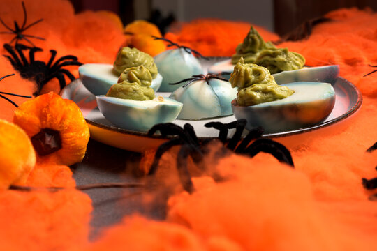 Deviled eggs decorated with green guacamole filling and black spiders on a plate surrounded by orange cobwebs and decorative pumpkins