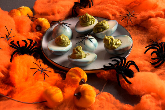 Deviled eggs decorated with green guacamole filling and black spiders on a plate surrounded by orange cobwebs and decorative pumpkins