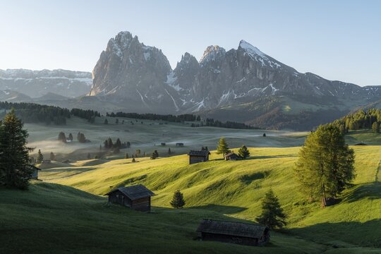 Hay barn and huts on misty meadows, behind Sassolungo and the Plattkofel, Alpe di Siusi, South Tyrol, Italy