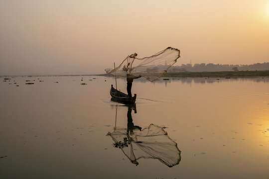 Fisherman with boat on Taung Tha Man Lake casts a fishing net for sunrise, reflection in the water, Thaung Tha Man Lake, Mandalay, Myanmar