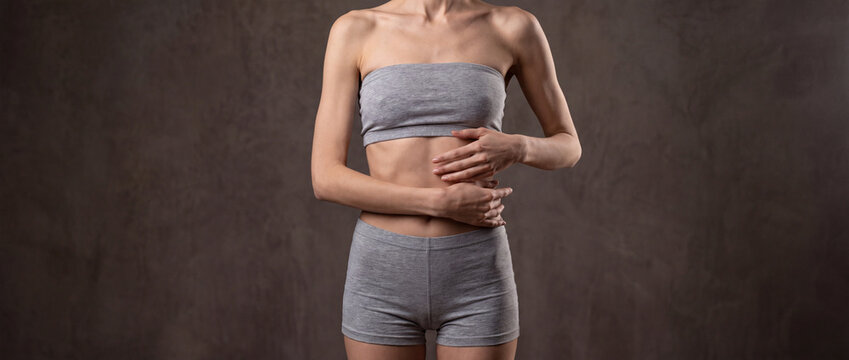 Skinny woman holding her bare stomach in grey underwear against a dark textured background, illustrating eating disorders, anorexia awareness, weight loss, and digestion issues