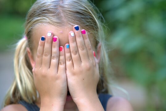 Blonde girl covers her face with both hands, bright pink and blue nail polish on her fingers, a green garden blurred behind her on a summer day