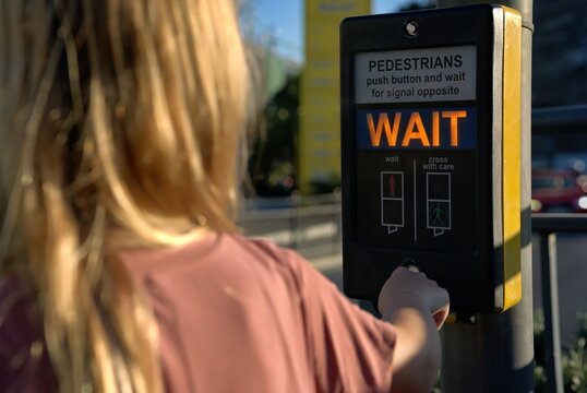 Girl in a pink shirt pushes the button of a pedestrian crossing box, the orange WAIT sign glowing in front of her, a street with cars blurred in the background