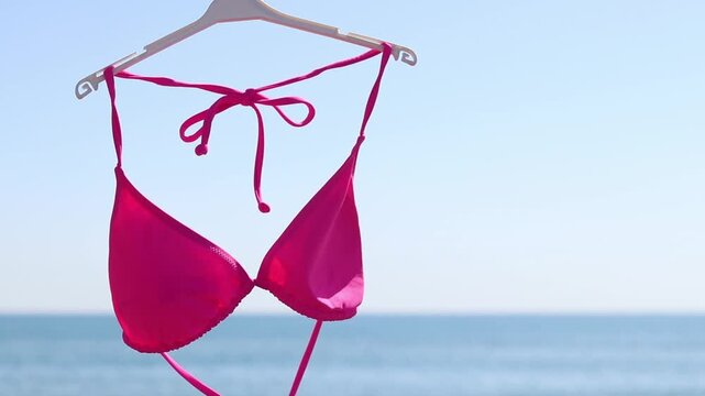 Bright pink bikini top hanging on a plastic hanger against a blurred blue sea and clear sky. Pink bikini close-up against the ocean on a sunny day. Concept of seaside resort, vacation