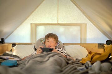Fototapeta premium Teenage boy lies under a blanket in a glamping tent and holds a phone with both hands, focused on a mobile game, soft daylight through the canvas walls
