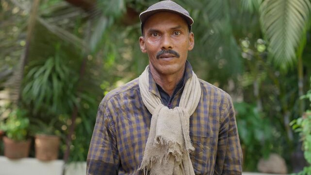 Daylight outdoor portrait shot of middle aged Indian Asian local male farmer wearing a cap and standing in the midst of a lush green tropical setting in the Konkan region, looking at the camera