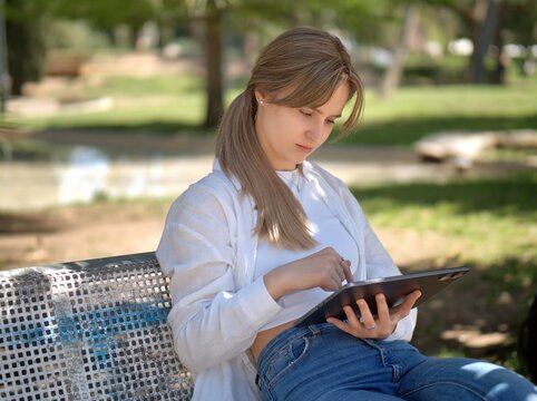 Young student scrolls through a tablet held on her lap while sitting on a metal park bench in the warm shade of the trees