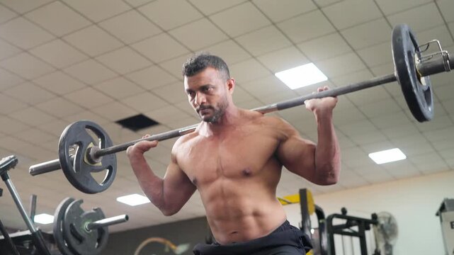 Muscular Indian Asian shirtless muscular middle aged man performing weighted barbell squats during strength or resistance training session in modern gym. Health, fitness, discipline, hard work concept
