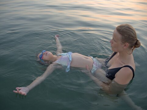 Woman in a black swimsuit holds her daughter on the surface of the calm sea, the child stretching her arms out as she floats