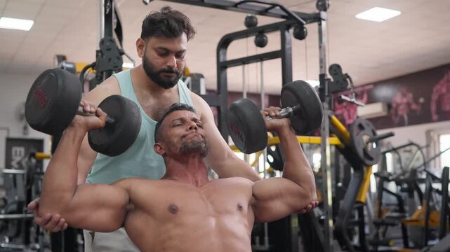 A muscular Indian Asian fit middle aged man performing a seated dumbbell press exercise with a colleague's support and supervision in a gym. Concept of teamwork, strength training, fitness, health