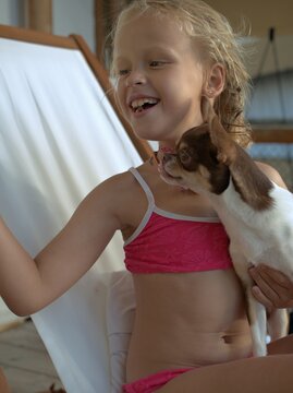 Girl laughs with her head thrown back, while a chihuahua stands up on her lap and leans against her shoulder in a canvas deck chair