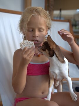 Girl holds a puffed rice cake to her mouth while a chihuahua on her lap sniffs at it with one paw lifted, both sitting in a canvas deck chair