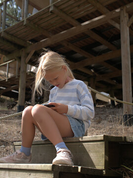 Blonde girl sits on wooden steps in shade holding a handheld game console, legs crossed