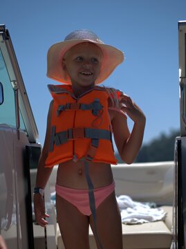Girl in a straw sun hat and an orange life jacket stands on a moving boat, touching her hair and laughing, blue sky and a green riverbank behind her