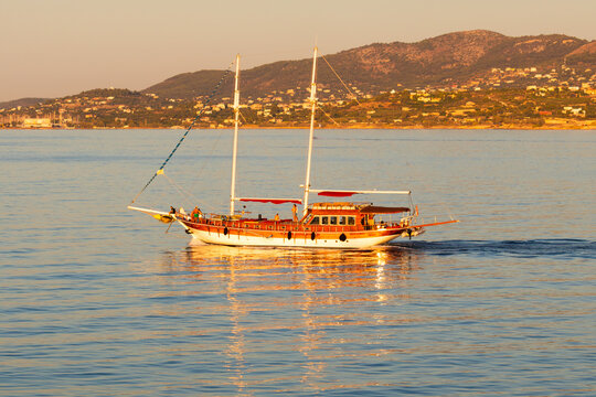 Traditional wooden sailing yacht cruising on calm sea waters at golden hour, with a scenic Greek coastal village in the background.