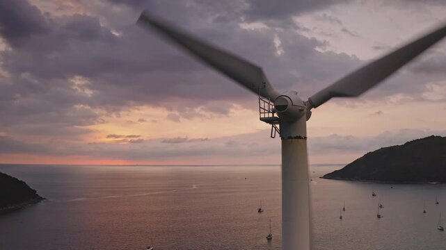 Close up wind turbine blades spinning, creating clean and sustainable energy high above the sea and coastline during a vibrant pink sunset over Phuket island, with boats dotting the water. Aerial shot