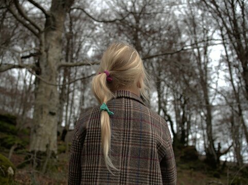 A girl with a colorful hair tie ponytail stands facing away in a tweed plaid coat among bare winter forest trees