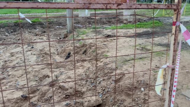 Fenced muddy pit with exposed roots and broken pavement. Red-and-white tape marks hazardous ground. Scene hints at recent flood or construction damage. Footage suitable for disaster, repair