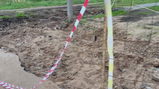 Fenced muddy pit with exposed roots and broken pavement. Red-and-white tape marks hazardous ground. Scene hints at recent flood or construction damage. Footage suitable for disaster, repair