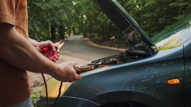 Driver stands in front of a car with an open hood, studying the battery terminals before connecting jumper cables. He tries to identify the positive and negative connections safely.