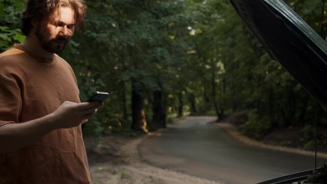 Driver ends a phone call beside a broken-down car with an open hood on a rural roadside. He now waits for a tow truck after requesting assistance.