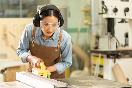 Female carpenter using table saw with safety gear in woodworking workshop