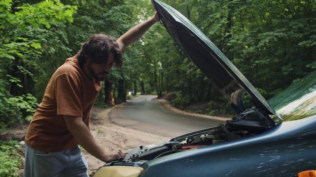 Driver holds the hood open and examines the engine bay of a broken-down car on a rural roadside. He assesses the mechanical problem before help arrives.