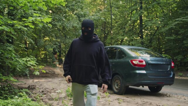 Masked thief hands car keys to buyer beside stolen vehicle in remote forest area. Illegal roadside deal shows transfer of stolen property and criminal deception.