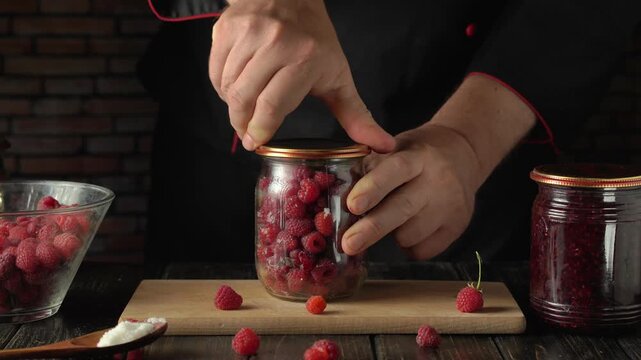 Hands of a male chef in black attire sealing a jar filled with fresh raspberries, with additional raspberries scattered on a wooden board and glass bowls in a rustic kitchen setting