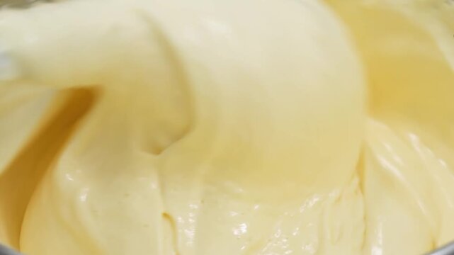 close-up, high-angle shot capturing a pastry chef wearing protective gloves as they vigorously whisk a smooth, thick, and creamy yellow vanilla custard or pudding mixture inside a stainless steel mixi
