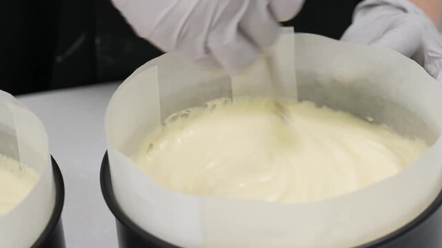 Close-up view of a pastry chef wearing white gloves carefully filling a round springform cake pan lined with high parchment paper collars. The smooth, velvety yellow batter or cream mixture is being p