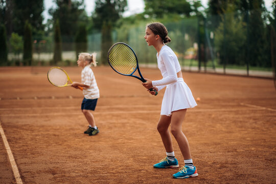 Two children practice tennis on an outdoor clay court, holding rackets in their hands and concentrating on a sports training exercise.