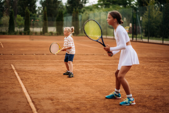 Two children practice tennis on an outdoor clay court, holding rackets in their hands and concentrating on a sports training exercise.