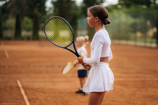 Two children practice tennis on an outdoor clay court, holding rackets in their hands and concentrating on a sports training exercise.