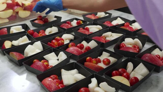 Close-up of a chef wearing blue gloves arranging fresh cut apples, pears, and cherry tomatoes into small black dessert containers on a marble countertop for catering or meal prep.