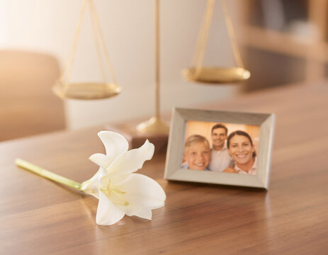 A solitary white flower rests on a wooden table; in the softly blurred background, a scale of justice and a family photo evoke the tragedy of a wrongful death.