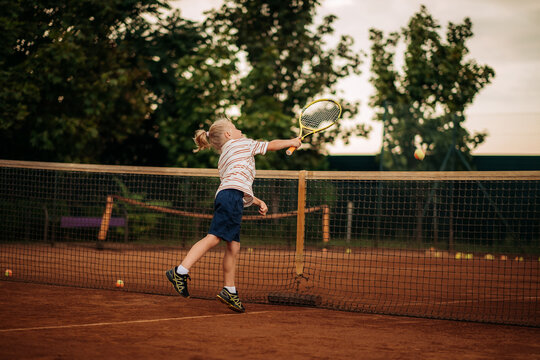 Little boy hitting tennis ball over net in jump on outdoor clay court, active childhood and dynamic sports training.