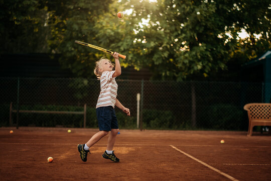 Little boy hitting tennis ball over net in jump on outdoor clay court, active childhood and dynamic sports training.