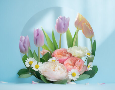 A pastel floral arrangement&mdash;featuring tulips, roses, and daisies amidst lush green leaves&mdash;is set against a soft blue background