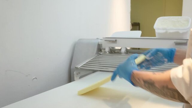Close-up shot of a professional baker wearing blue protective gloves, carefully guiding a flat block of pastry dough onto the conveyor belt of an industrial motorized dough sheeter machine to roll it 