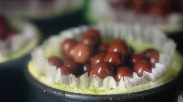 Macro footage of gourmet fruit tartlets in small baking pans during preparation. The focus highlights a collection of small, shiny chocolate spheres or dark berries resting inside a pleated white pape