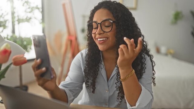 Woman holding smartphone and resting chin on fist while smiling and gesturing at screen in a bedroom; remote work engaged.